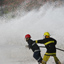 Two men in firefighting uniforms and hardhats holding a hose with spraying water, wall of foam spraying up in background