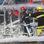 Two men in firefighting uniforms and hardhats holding a hose with water spraying into a burned-out bus
