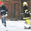 Two men in firefighting uniforms and hardhats standing in foam on ground, holding a hose beside the corner of a burned-out bus