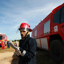 Man in firefighting uniform, red hardhat and brown facemask standing in front of a red truck, two men in uniforms and hardhats with red truck in background