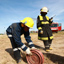Two men in firefighting uniforms and hardhats, one standing, one bent forward holding a rolled-up hose