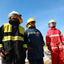 Three men in firefighting uniforms and hardhats standing, blue sky in backfround