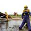 Three men in brown firefighting uniforms and yellow hardhats, one in front holding a dummy of a person, one behind holding hose spraying water at burned-out vehicle, one in background running away toward a red firetruck
