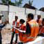 Man in black uniform and cap faces group of men and women in orange vests, all standing in front of white building with no roof or windows
