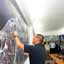 Man in black shirt points to a point on a large map hanging on wall in a room, people in background wath