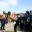 Shirtless man with back to camera kicks soldiers at clear shields while another observes