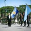 Man in black suit walks by row of uniformed soldiers and two blue flags