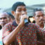 Man in colourful shirt speaks into a microphone to an unseen person while two men stand behind him