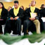 Three men and one woman all wearing black robes and yellow shirts sit watching an unseen speaker