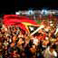 Unseen individual waves a large colourful flag amid a large crowd of people at night
